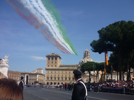 Jets fly overhead releasing smoke trails in the colors of the Italian flag against a clear blue sky, with an ornate historic building and a dome in the background. Trees and a crowd of people are visible, along with uniformed personnel in the foreground.