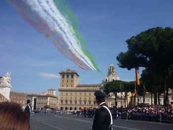 Jets fly overhead releasing smoke trails in the colors of the Italian flag against a clear blue sky, with an ornate historic building and a dome in the background. Trees and a crowd of people are visible, along with uniformed personnel in the foreground.