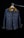 A denim jacket hangs on a wooden hanger against a dark background. The jacket features a brown collar, two front flap pockets with metal buttons, and detailed stitching. The lighting creates a focus on the texture of the fabric and the structure of the garment.