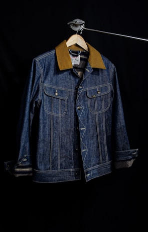 A denim jacket hangs on a wooden hanger against a dark background. The jacket features a brown collar, two front flap pockets with metal buttons, and detailed stitching. The lighting creates a focus on the texture of the fabric and the structure of the garment.