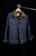 A denim jacket hangs on a wooden hanger against a dark background. The jacket features a brown collar, two front flap pockets with metal buttons, and detailed stitching. The lighting creates a focus on the texture of the fabric and the structure of the garment.