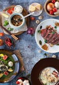 A flat lay of various dishes and ingredients on a textured surface. In the center, there is a bowl with a soup-like dish accompanied by a spoon and a side of rice. A plate with roasted meat, garnished with vegetables and breadcrumbs, is beside it. Fresh ingredients like cheese, mushrooms, tomatoes, onions, and herbs are spread around. The overall presentation is artistic and vibrant.
