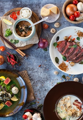A flat lay of various dishes and ingredients on a textured surface. In the center, there is a bowl with a soup-like dish accompanied by a spoon and a side of rice. A plate with roasted meat, garnished with vegetables and breadcrumbs, is beside it. Fresh ingredients like cheese, mushrooms, tomatoes, onions, and herbs are spread around. The overall presentation is artistic and vibrant.
