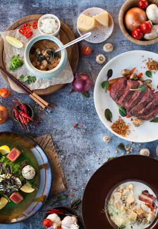A flat lay of various dishes and ingredients on a textured surface. In the center, there is a bowl with a soup-like dish accompanied by a spoon and a side of rice. A plate with roasted meat, garnished with vegetables and breadcrumbs, is beside it. Fresh ingredients like cheese, mushrooms, tomatoes, onions, and herbs are spread around. The overall presentation is artistic and vibrant.