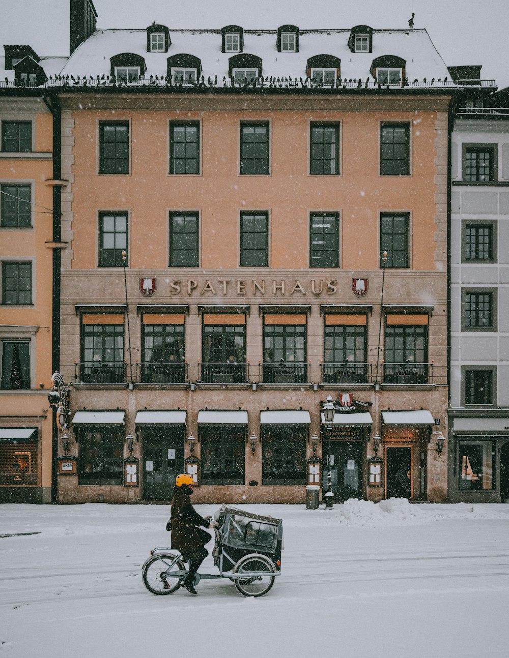낮 동안 갈색 콘크리트 건물 근처의 눈 덮인 도로에서 검은 오토바이를 타는 사람