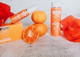 A set of orange-themed beauty products, including a soap box and a bottle labeled 'Brightening C+ with Glutathione', is surrounded by fresh oranges and red petals. The scene is set against a textured light background, creating a vibrant and fresh atmosphere.