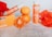 A set of orange-themed beauty products, including a soap box and a bottle labeled 'Brightening C+ with Glutathione', is surrounded by fresh oranges and red petals. The scene is set against a textured light background, creating a vibrant and fresh atmosphere.