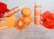 A set of orange-themed beauty products, including a soap box and a bottle labeled 'Brightening C+ with Glutathione', is surrounded by fresh oranges and red petals. The scene is set against a textured light background, creating a vibrant and fresh atmosphere.