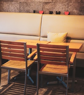 A cozy cafe setting with a wooden table and two matching chairs in the foreground. Behind the table, a cushioned bench lines the wall, adorned with two red heart shapes and black letters. The floor has a rustic concrete design, and soft, warm lighting creates a relaxed atmosphere.