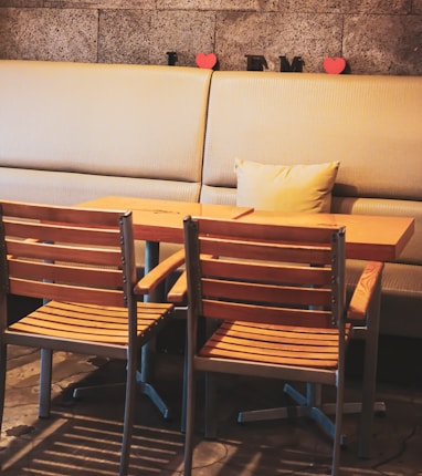 A cozy cafe setting with a wooden table and two matching chairs in the foreground. Behind the table, a cushioned bench lines the wall, adorned with two red heart shapes and black letters. The floor has a rustic concrete design, and soft, warm lighting creates a relaxed atmosphere.