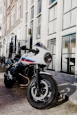 A sleek, white motorcycle with red and blue stripes is parked on a street lined with tall, elegant buildings featuring large windows. The motorcycle's design is modern and sporty, with a round headlight and detailed, sturdy tires. The surrounding area appears to be an urban setting, with the street and buildings casting shadows on the pavement.