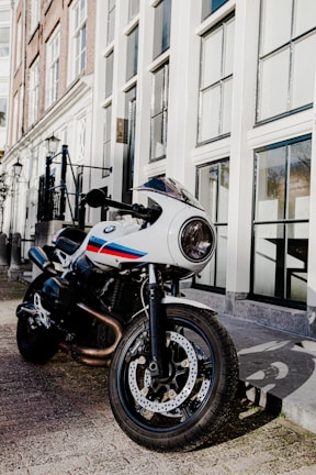 A sleek, white motorcycle with red and blue stripes is parked on a street lined with tall, elegant buildings featuring large windows. The motorcycle's design is modern and sporty, with a round headlight and detailed, sturdy tires. The surrounding area appears to be an urban setting, with the street and buildings casting shadows on the pavement.