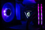 A computer interior featuring bright LED lighting and components. A large cooling fan emits a deep blue light on the left side. In the center, an illuminated logo with a dragon design is visible on a component. Two vertical memory sticks on the right side emit a vivid pink glow. The overall scene emphasizes the vibrant and high-tech aesthetics of modern PC builds.