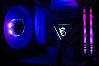 A computer interior featuring bright LED lighting and components. A large cooling fan emits a deep blue light on the left side. In the center, an illuminated logo with a dragon design is visible on a component. Two vertical memory sticks on the right side emit a vivid pink glow. The overall scene emphasizes the vibrant and high-tech aesthetics of modern PC builds.
