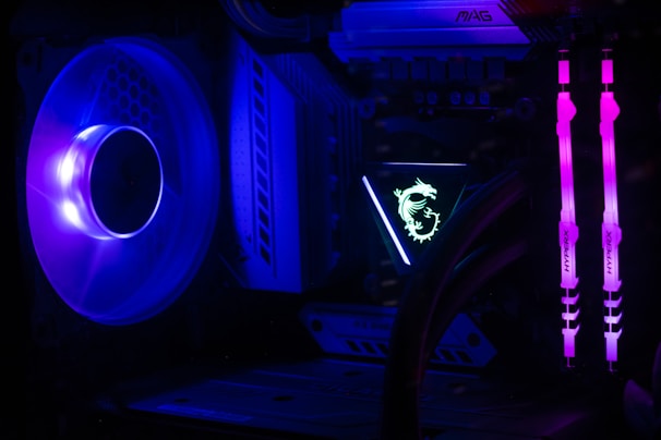A computer interior featuring bright LED lighting and components. A large cooling fan emits a deep blue light on the left side. In the center, an illuminated logo with a dragon design is visible on a component. Two vertical memory sticks on the right side emit a vivid pink glow. The overall scene emphasizes the vibrant and high-tech aesthetics of modern PC builds.