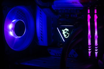 A computer interior featuring bright LED lighting and components. A large cooling fan emits a deep blue light on the left side. In the center, an illuminated logo with a dragon design is visible on a component. Two vertical memory sticks on the right side emit a vivid pink glow. The overall scene emphasizes the vibrant and high-tech aesthetics of modern PC builds.