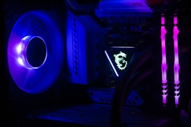 A computer interior featuring bright LED lighting and components. A large cooling fan emits a deep blue light on the left side. In the center, an illuminated logo with a dragon design is visible on a component. Two vertical memory sticks on the right side emit a vivid pink glow. The overall scene emphasizes the vibrant and high-tech aesthetics of modern PC builds.