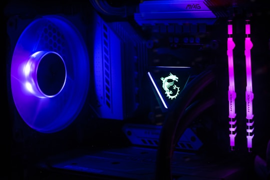A computer interior featuring bright LED lighting and components. A large cooling fan emits a deep blue light on the left side. In the center, an illuminated logo with a dragon design is visible on a component. Two vertical memory sticks on the right side emit a vivid pink glow. The overall scene emphasizes the vibrant and high-tech aesthetics of modern PC builds.