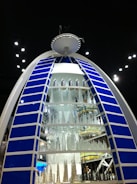 A large, illuminated structure resembling a futuristic building display with a semi-circular shape and multiple levels of miniature skyscraper models made of glass or a reflective material. The structure is framed by blue panels with white borders, and it stands out against a dark background with scattered lights.