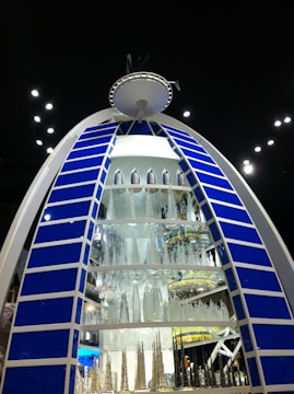 A large, illuminated structure resembling a futuristic building display with a semi-circular shape and multiple levels of miniature skyscraper models made of glass or a reflective material. The structure is framed by blue panels with white borders, and it stands out against a dark background with scattered lights.