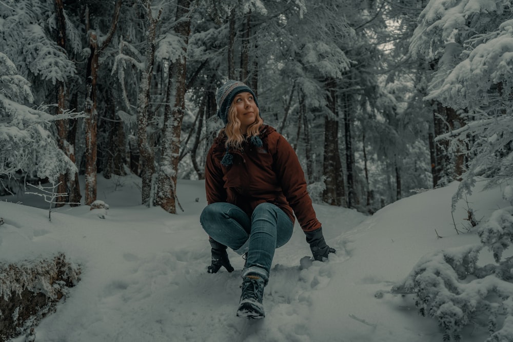 donna in giacca marrone e jeans blu denim seduta su terreno coperto di neve