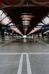 A symmetrical and industrial train platform under construction, featuring metal beams and a netted ceiling. The tracks extend into the distance, flanked by platforms with minimal human activity. Station signs and clocks are visible, adding to the utilitarian atmosphere.