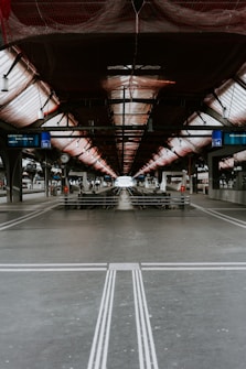 A symmetrical and industrial train platform under construction, featuring metal beams and a netted ceiling. The tracks extend into the distance, flanked by platforms with minimal human activity. Station signs and clocks are visible, adding to the utilitarian atmosphere.