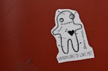 A sticker on a reddish-brown surface features a stylized cartoon voodoo doll with a stitched mouth and eyes. The doll has a large black heart on its chest and pins sticking out from its body. The sticker includes the text '@molly-sandra_ Voodoo like to love me?'