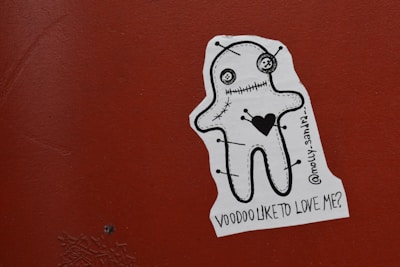 A sticker on a reddish-brown surface features a stylized cartoon voodoo doll with a stitched mouth and eyes. The doll has a large black heart on its chest and pins sticking out from its body. The sticker includes the text '@molly-sandra_ Voodoo like to love me?'
