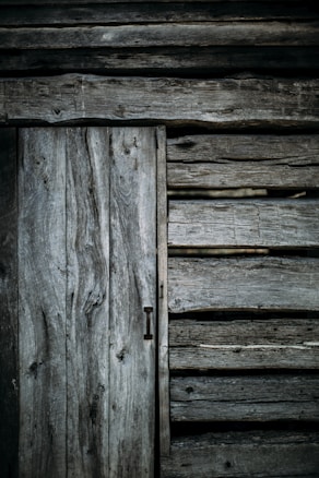 Weathered wooden planks form the exterior of a rustic, aged structure. The texture of the wood appears rough and worn, with visible knots and grain patterns, contributing to an overall antique and rugged look.