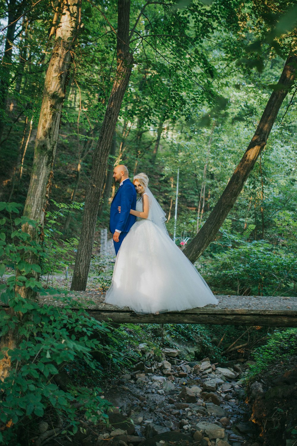 bride and groom standing on brown wooden bridge surrounded by green trees during daytime