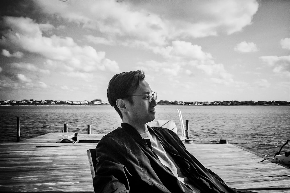 grayscale photo of woman in black jacket sitting on wooden dock