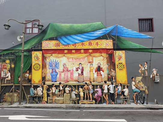 A painted mural depicting a traditional Chinese opera performance. The setting includes vibrant costumes worn by the actors on stage, surrounded by an audience of people dressed in casual attire. The background features decorative elements like intricate Chinese motifs and a green and blue fabric canopy. Street elements like a lamppost and a cart with a basket of goods are also present.