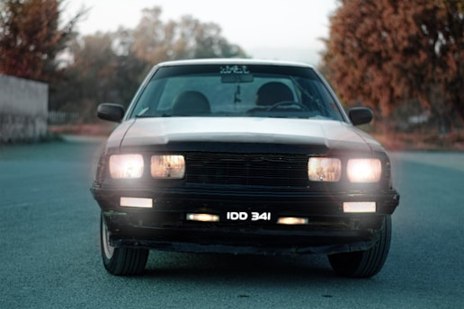 A vintage car with headlights on is positioned on a deserted road, surrounded by blurred foliage in the background with a soft focus. The license plate reads 'IDD 341', and the car's classic design features a prominent grille and wide headlights.