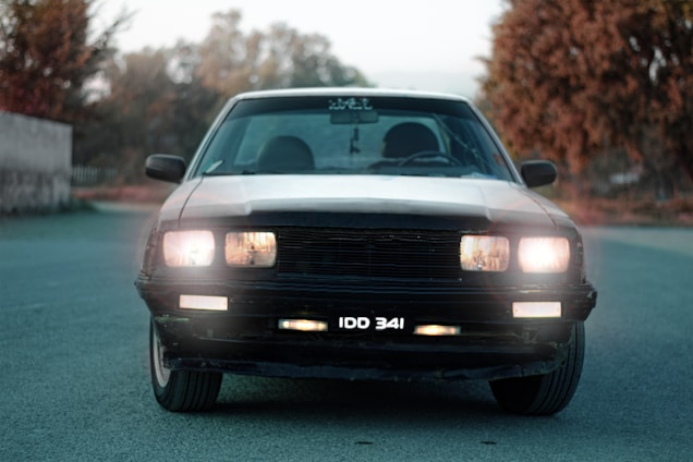 A vintage car with headlights on is positioned on a deserted road, surrounded by blurred foliage in the background with a soft focus. The license plate reads 'IDD 341', and the car's classic design features a prominent grille and wide headlights.