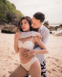 A couple is posing intimately on a sandy beach. The woman is wearing a white swimsuit, and the man is in a striped shirt and patterned shorts. The beach has rocky outcrops and lush green foliage in the background under a bright sky.