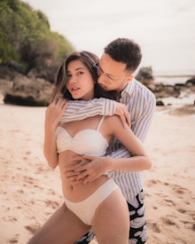 A couple is posing intimately on a sandy beach. The woman is wearing a white swimsuit, and the man is in a striped shirt and patterned shorts. The beach has rocky outcrops and lush green foliage in the background under a bright sky.