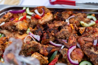 A close-up view of grilled chicken pieces mixed with sliced cucumbers, red onions, and red chili peppers on a serving platter. The meat is well-cooked with visible charring, indicating it has been grilled or roasted. The vegetables add a fresh contrast to the rich texture of the meat.