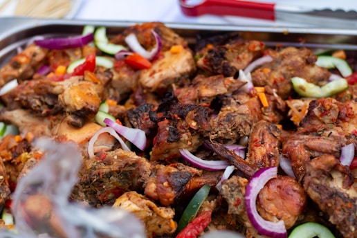 A close-up view of grilled chicken pieces mixed with sliced cucumbers, red onions, and red chili peppers on a serving platter. The meat is well-cooked with visible charring, indicating it has been grilled or roasted. The vegetables add a fresh contrast to the rich texture of the meat.