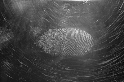 A black and white image featuring a prominent fingerprint on a surface with scratch marks. The texture and details of the fingerprint are clearly visible, suggesting close-up photography.