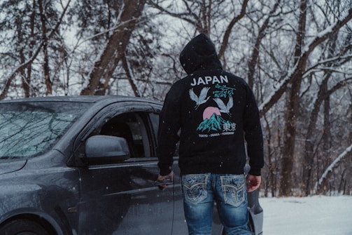 A person wearing a hooded sweatshirt featuring Japanese motifs is standing next to a dark-colored car in a snowy forest. The scene is framed by bare trees covered with snow, with a grayish sky visible in the background. The sweatshirt has a design of cranes and mountains, accompanied by Japanese text.