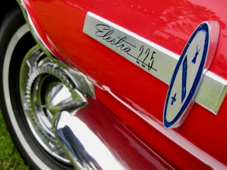A close-up of a red car with a shiny chrome trim featuring the text 'Electra 225'. The image shows a part of a classic car's body including a stylized logo.