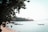 A serene beach scene with calm blue waters and several colorful longtail boats floating nearby. Lush green foliage overhangs from the left, and in the distance, a small island or coastal landmass with scattered palm trees is visible under a clear sky.