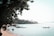 A serene beach scene with calm blue waters and several colorful longtail boats floating nearby. Lush green foliage overhangs from the left, and in the distance, a small island or coastal landmass with scattered palm trees is visible under a clear sky.