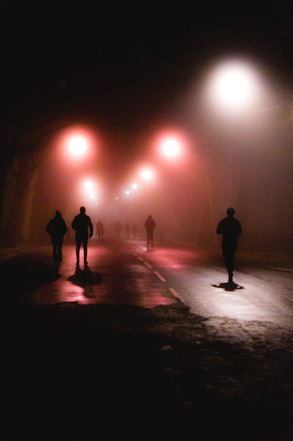 Silhouettes of people moving along a foggy, lamplit street at night.