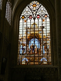 A large stained glass window depicts a religious scene with figures in robes. The intricate design features vibrant colors and detailed patterns, surrounded by gothic-style architecture. Crest-like symbols are displayed above the figures.