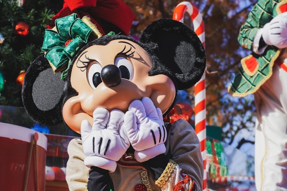 A large costumed character with black ears and a red bow is covering its face with its gloved hands. The character stands in an outdoor setting decorated with festive elements, including a Christmas tree and candy cane. Bright sunlight enhances the vibrant colors and cheerful atmosphere.
