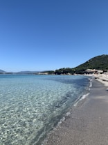 A serene beach scene with crystal-clear, turquoise water gently lapping against a sandy shore. The sky is a clear, deep blue, with a lush green hill to the right. A few scattered buildings are visible in the distance, nestled among the greenery.
