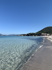 A serene beach scene with crystal-clear, turquoise water gently lapping against a sandy shore. The sky is a clear, deep blue, with a lush green hill to the right. A few scattered buildings are visible in the distance, nestled among the greenery.