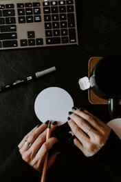A close-up view of a workspace featuring a keyboard, a cup, and a small notepad or coasters with a person's hands writing or drawing on it. The hand holds a pencil and a pen, and a marker is nearby.